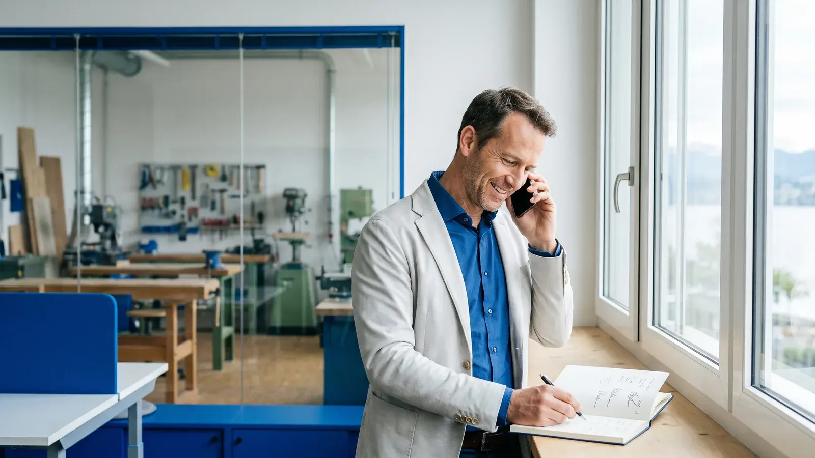 Unternehmer telefoniert in seinem hellen Büro neben der Werkstatt und macht sich Notizen