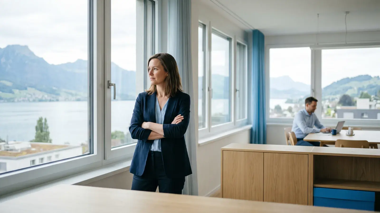 Gründerin steht nachdenklich mit verschränkten Armen am Bürofenster, im Hintergrund ein Kollege am Tisch