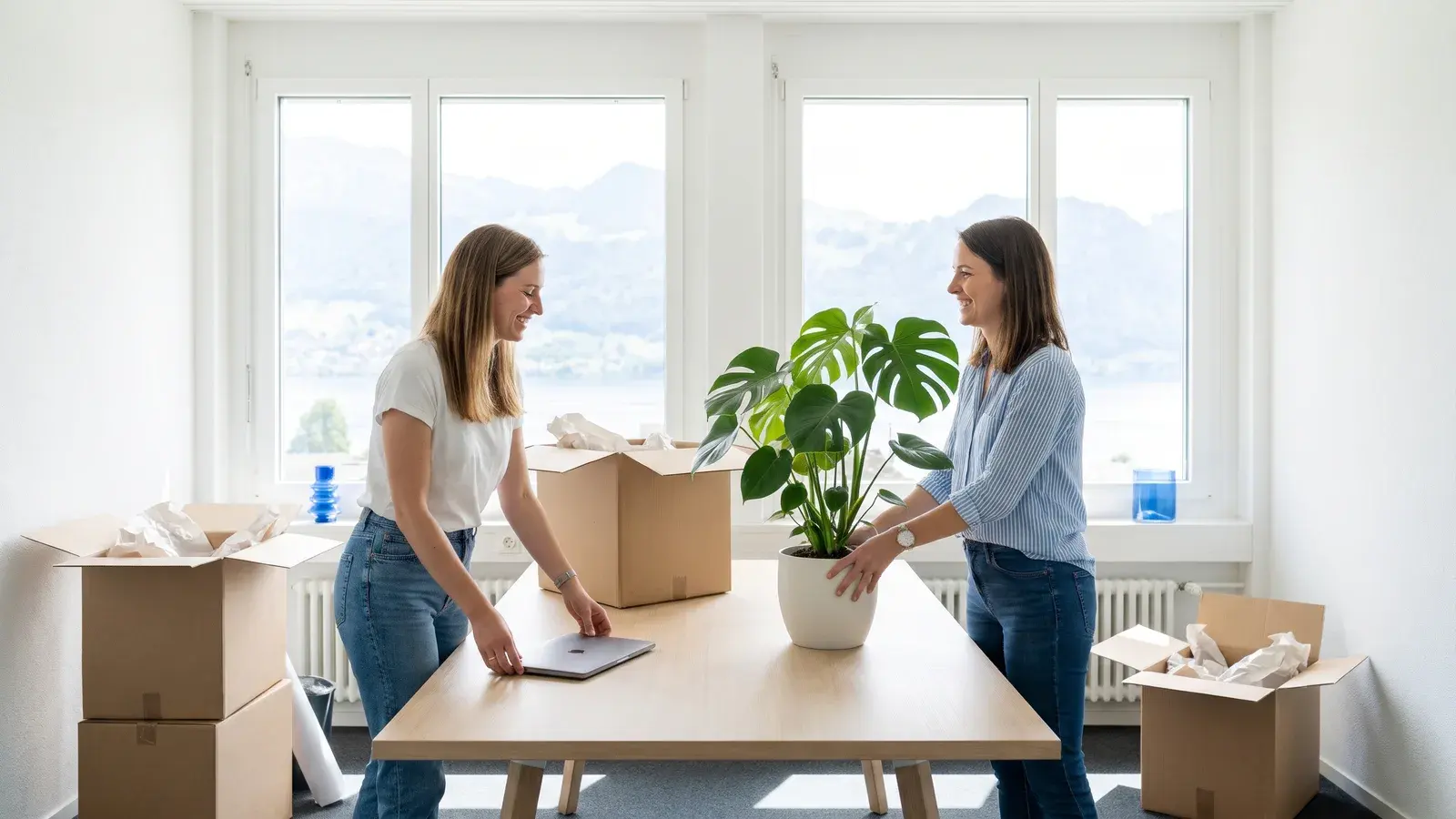 Zwei Gründerinnen packen in einem frisch bezogenen, hellen Büro Kartons aus und stellen einen zugeklappten Laptop auf den neuen Tisch